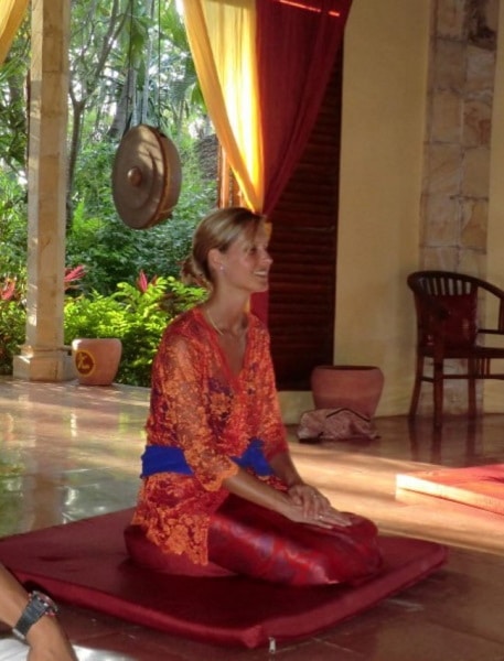 Aurélie Thomas assise sur un coussin de méditation, vêtue de façon traditionnelle à Bali, les mains posées sur ses cuisses, les yeux fermés et le sourire aux lèvres