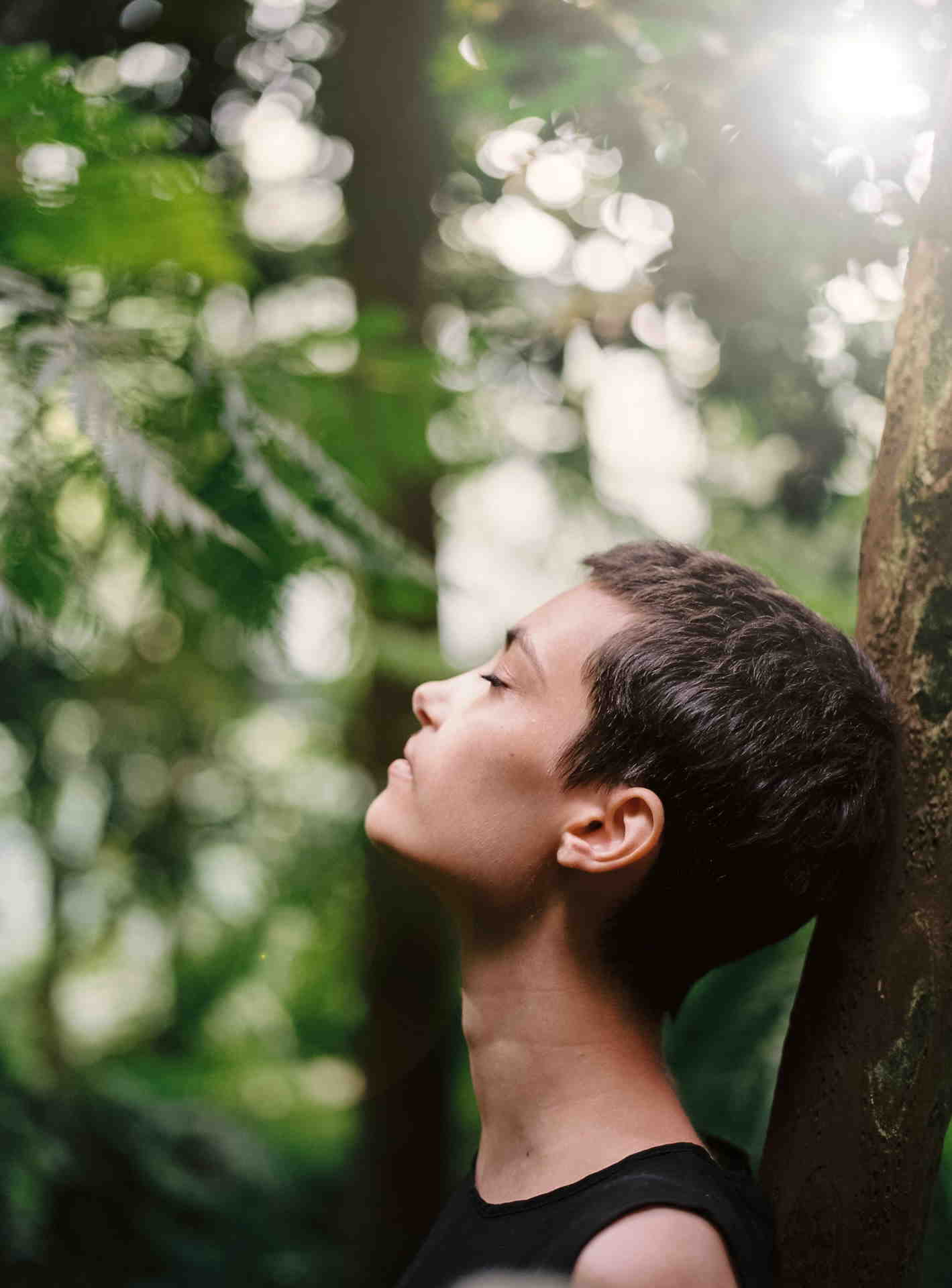 femme de profil aux cheveux courts, les yeux fermés, qui appuie sa tête de dos contre le tronc d'un arbre en forêt