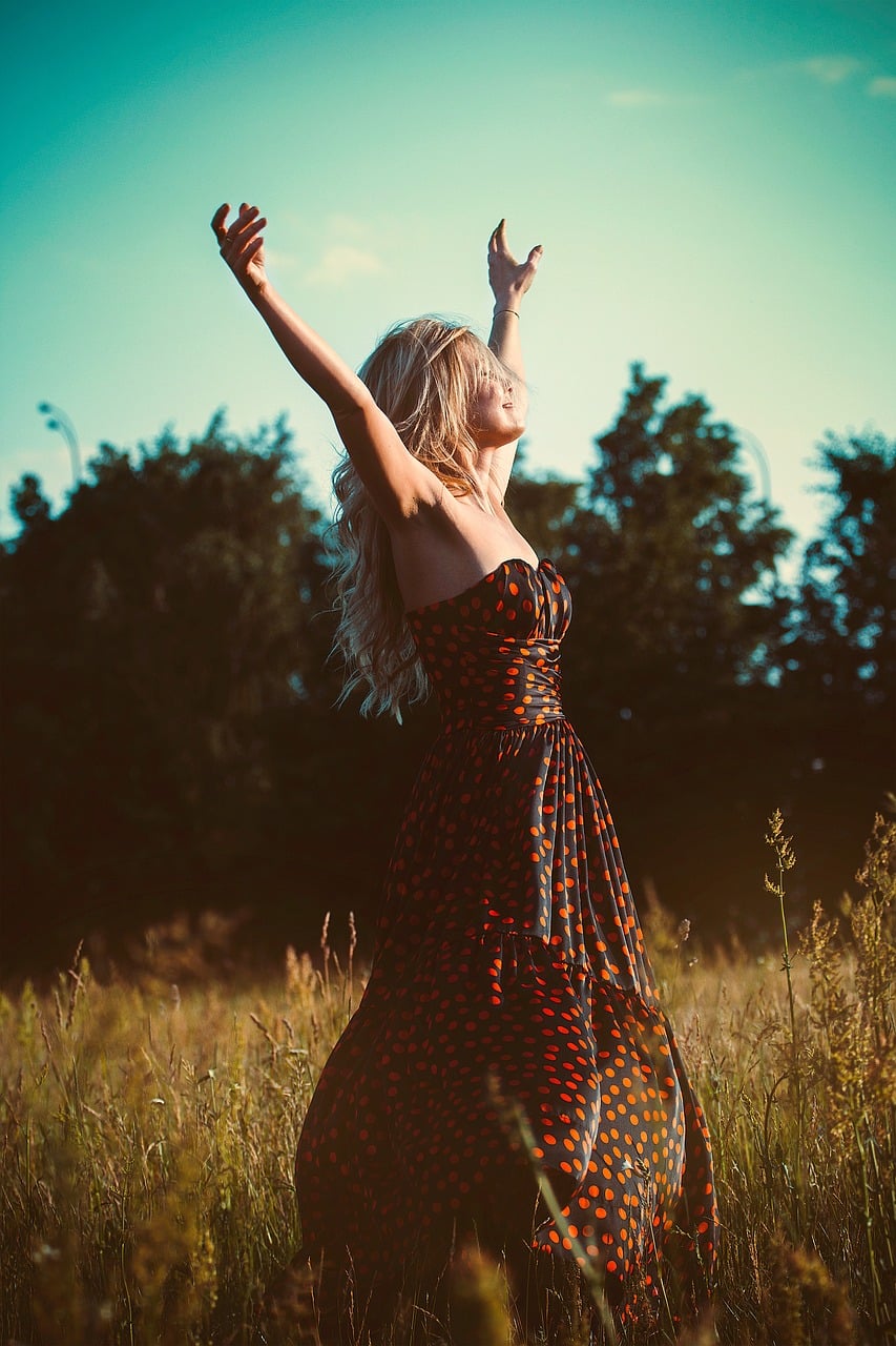 femme debout dans une prairie, vêtue d'une robe noire à pois rouges, qui écarte les bras tendus en l'air vers le ciel, le sourire aux lèvres