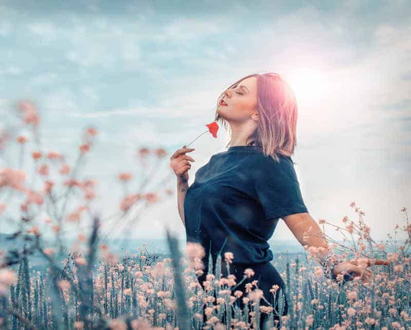 femme souriante dans une prairie portant un coquelicot vers son menton, les yeux fermés