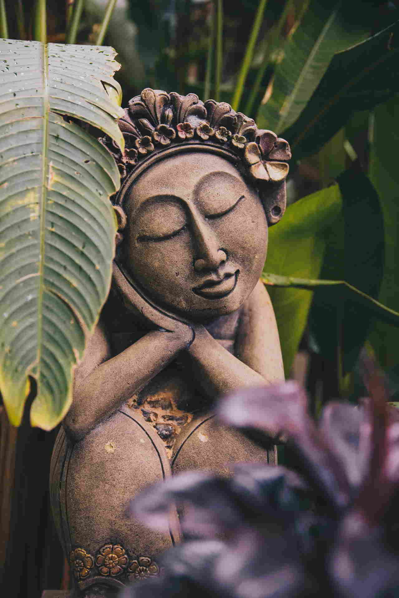 statuette de femme balinaise en bois, les yeux fermés, ses mains jointes sont accolées sur sa joue droite, avec une couronne de fleurs sculptée, posée au milieu d'une végétation luxuriante